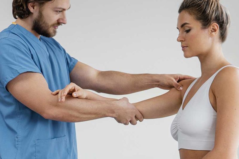A male chiropractor in blue scrubs examines a woman's arm, who wears a comfortable, flexible white sports bra.